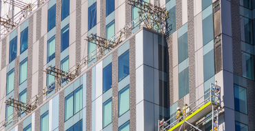 Workers on a suspended scaffolding platform conducting maintenance on the facade of a modern glass office building with patterned exterior, showcasing urban architecture and construction safety practices in a city environment.