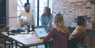 Team meeting in a modern office space with exposed brick walls, featuring two men and two women discussing work. One man stands addressing the group while the others are seated at a wooden table with laptops and office supplies, in a collaborative and casual business environment.